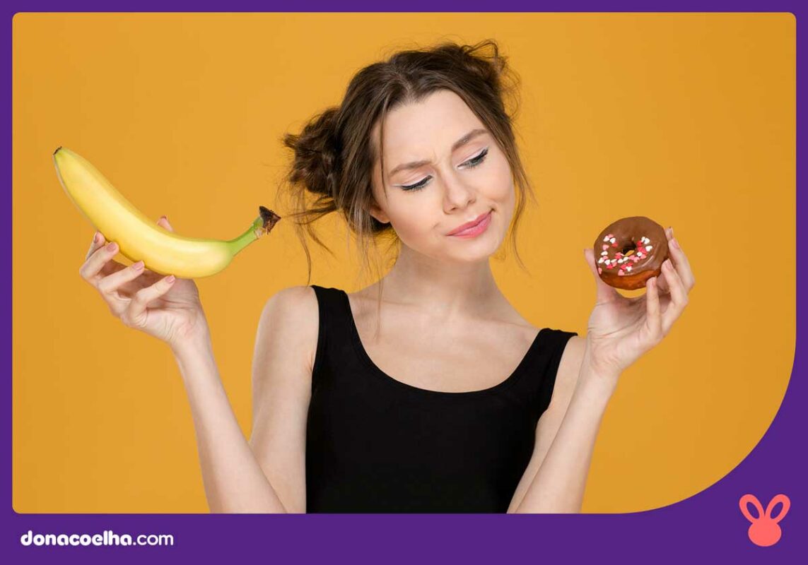 Pessoa segurando dois alimentos, sendo um uma banana e outro uma rosquina, ao qual ela está olhando